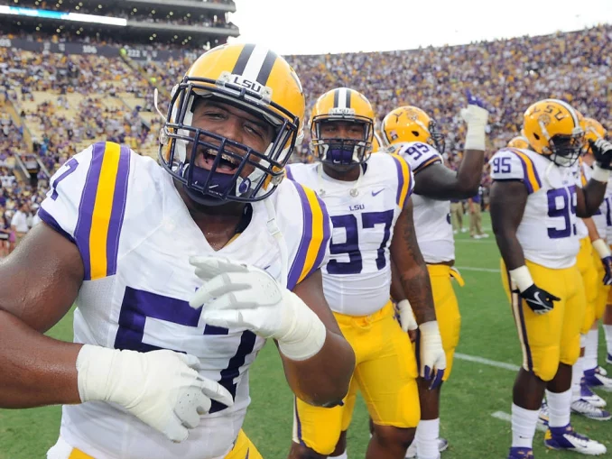 LSU football player in white jersey and yellow pants (#7) celebrating on the field with teammates; crowded stadium in background.