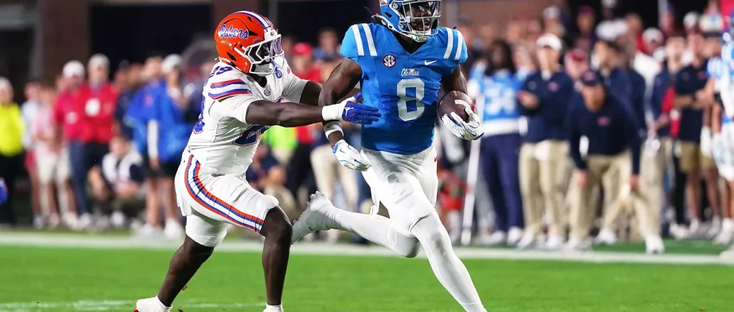 Ole Miss running back in blue carries the ball as a Florida defender in white chases him on a green field.