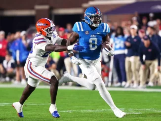 Ole Miss running back in blue carries the ball as a Florida defender in white chases him on a green field.