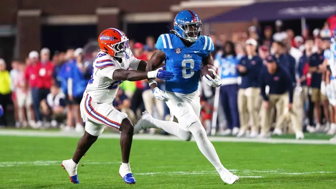 Ole Miss running back in blue carries the ball as a Florida defender in white chases him on a green field.