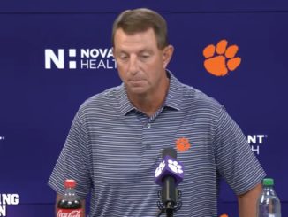 Man at a podium during a press conference, wearing a striped polo with a Clemson paw logo; backdrop shows Clemson branding and microphones on the table.