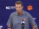 Man at a podium during a press conference, wearing a striped polo with a Clemson paw logo; backdrop shows Clemson branding and microphones on the table.