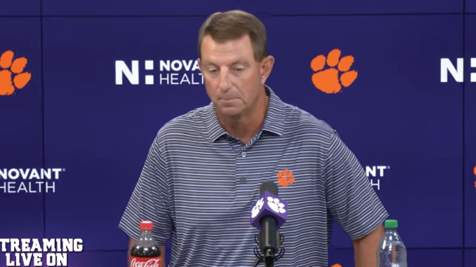 Man at a podium during a press conference, wearing a striped polo with a Clemson paw logo; backdrop shows Clemson branding and microphones on the table.