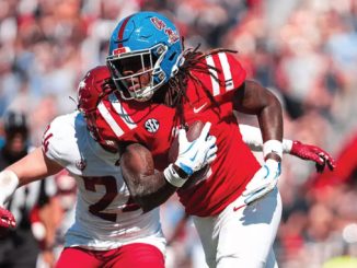 College football player in red uniform runs with the ball as a defender in white closes in for a tackle; SEC patch visible on the opponent's jersey.