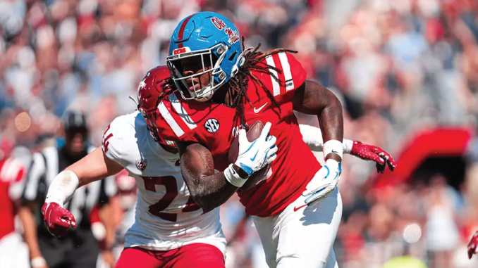College football player in red uniform runs with the ball as a defender in white closes in for a tackle; SEC patch visible on the opponent's jersey.