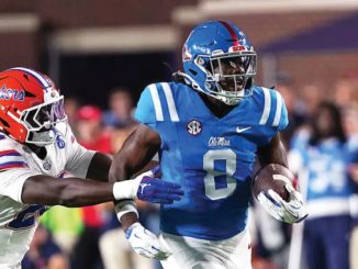 Ole Miss Rebels player in blue jersey carries the football, pursued by a Florida Gators defender in white.