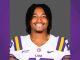 Portrait of a smiling LSU football player in a white jersey with purple and gold stripes, standing against a purple background.