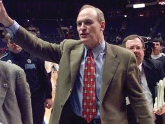Man in a brown blazer waves to the crowd on a basketball court, with reporters and security visible in the background.