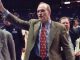 Man in a brown blazer waves to the crowd on a basketball court, with reporters and security visible in the background.