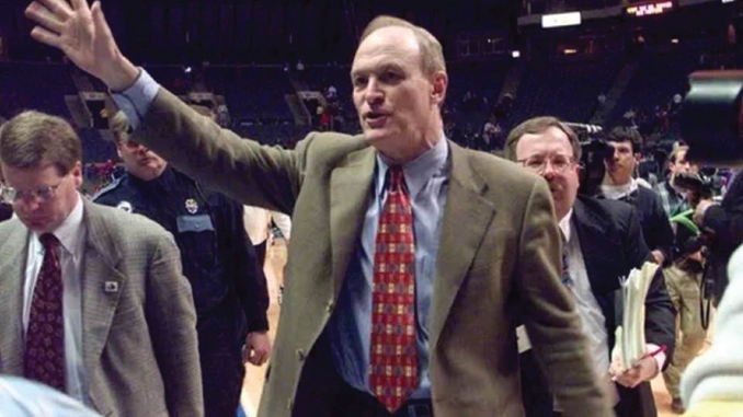 Man in a brown blazer waves to the crowd on a basketball court, with reporters and security visible in the background.