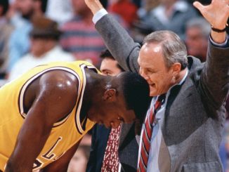 Basketball coach with arms raised in celebration beside a kneeling Lakers player in a yellow jersey, crowd in background.
