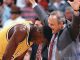Basketball coach with arms raised in celebration beside a kneeling Lakers player in a yellow jersey, crowd in background.