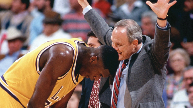 Basketball coach with arms raised in celebration beside a kneeling Lakers player in a yellow jersey, crowd in background.