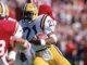 LSU football running back wearing #21 in white and gold jersey carries the ball as a defender in red closes in.