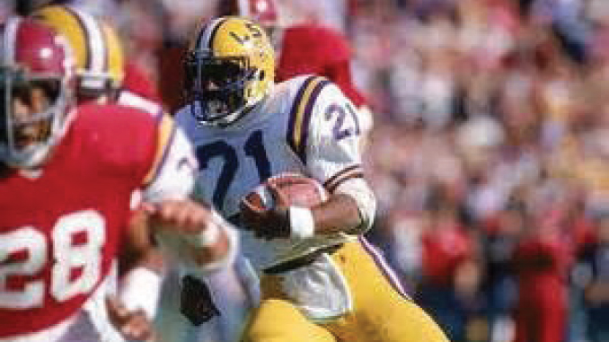 LSU football running back wearing #21 in white and gold jersey carries the ball as a defender in red closes in.