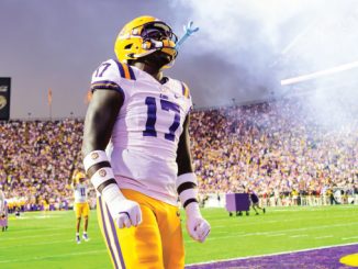 LSU football player wearing number 17 badge stands on the field amid a cheering stadium and smoke from pyrotechnics in the background.