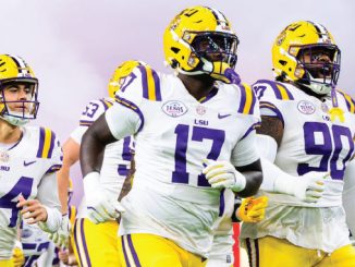 LSU Tigers football players in white uniforms run onto the field wearing purple and yellow helmets.