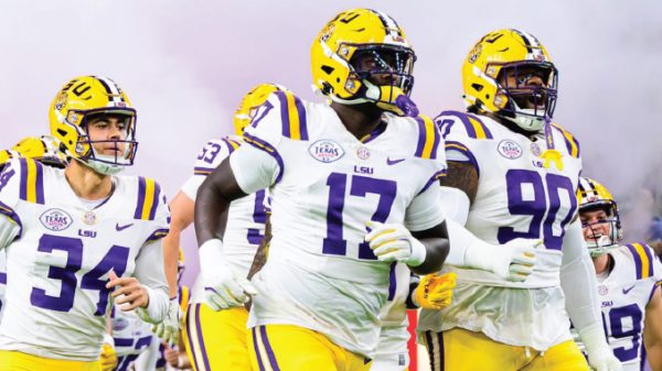 LSU Tigers football players in white uniforms run onto the field wearing purple and yellow helmets.