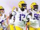 LSU Tigers football players in white uniforms run onto the field wearing purple and yellow helmets.