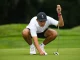 Golfer in a white polo and cap crouches on the green, lining up a putt with a putter near the ball.