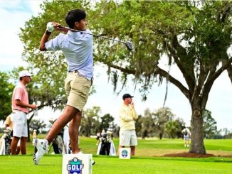 Golfer mid-swing on a tee with onlookers at a sunny golf course.