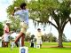 Golfer mid-swing on a tee with onlookers at a sunny golf course.