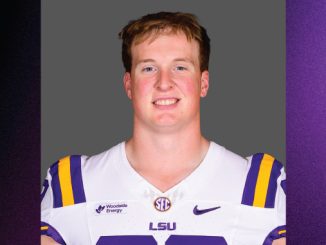 Portrait of a smiling male college football player in LSU white jersey with purple and gold stripes and shoulder pads, neutral gray background.