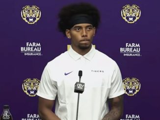 Athlete at a LSU press conference wearing a white polo, speaking into a microphone against a purple backdrop with logos.