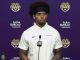 Athlete at a LSU press conference wearing a white polo, speaking into a microphone against a purple backdrop with logos.
