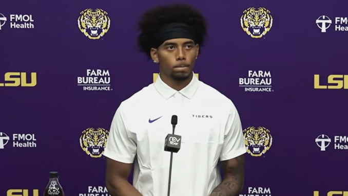 Athlete at a LSU press conference wearing a white polo, speaking into a microphone against a purple backdrop with logos.