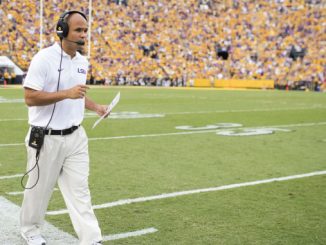 LSU football coach with headset and clipboard walking along the sideline during a game, yellow-clad crowd in the stands.