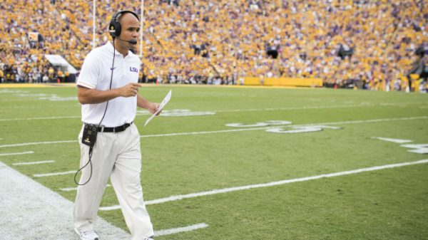 LSU football coach with headset and clipboard walking along the sideline during a game, yellow-clad crowd in the stands.