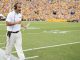 LSU football coach with headset and clipboard walking along the sideline during a game, yellow-clad crowd in the stands.