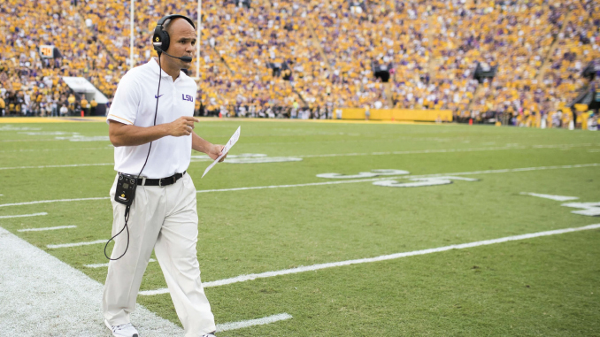 LSU football coach with headset and clipboard walking along the sideline during a game, yellow-clad crowd in the stands.