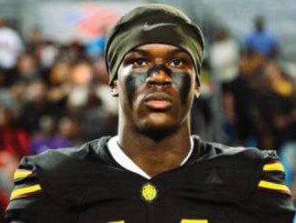 Football player in black jersey with yellow stripes and eye black, standing on the field with a blurred crowd in the background.
