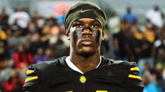 Football player in black jersey with yellow stripes and eye black, standing on the field with a blurred crowd in the background.