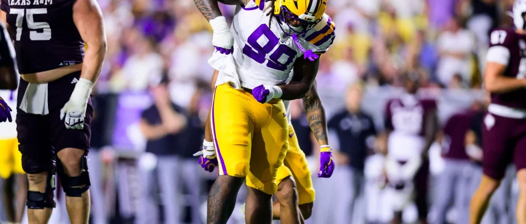 LSU football player in white jersey and gold pants collides with a Texas A&M defender during a college football game on a green field.