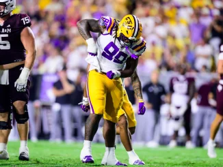 LSU football player in white jersey and gold pants collides with a Texas A&M defender during a college football game on a green field.