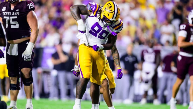 LSU football player in white jersey and gold pants collides with a Texas A&M defender during a college football game on a green field.