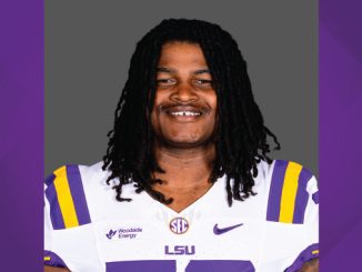 Portrait of a smiling LSU football player in a white jersey with purple and gold stripes and LSU logo, against a neutral backdrop.