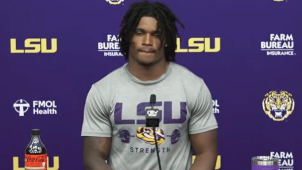 LSU football player at a press conference standing behind a microphone, wearing a gray 'LSU Strength' shirt against a purple LSU backdrop with logos and a Coke bottle on the table.