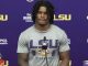 LSU football player at a press conference standing behind a microphone, wearing a gray 'LSU Strength' shirt against a purple LSU backdrop with logos and a Coke bottle on the table.