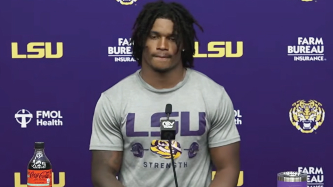 LSU football player at a press conference standing behind a microphone, wearing a gray 'LSU Strength' shirt against a purple LSU backdrop with logos and a Coke bottle on the table.