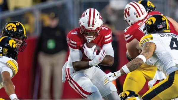 College football action: a player in a red jersey cradles the ball while defenders in black-and-yellow uniforms close in
