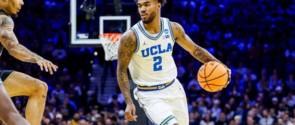 UCLA basketball guard in white jersey dribbling the ball during a game with a crowded arena in the background