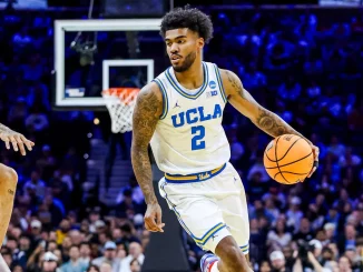 UCLA basketball guard in white jersey dribbling the ball during a game with a crowded arena in the background