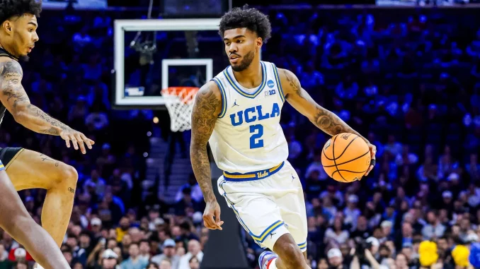 UCLA basketball guard in white jersey dribbling the ball during a game with a crowded arena in the background