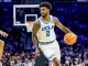 UCLA basketball guard in white jersey dribbling the ball during a game with a crowded arena in the background