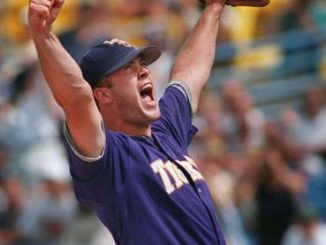 Baseball pitcher in a purple uniform raises his glove and shouts in celebration on the mound.