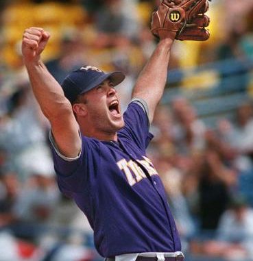 Baseball pitcher in a purple uniform raises his glove and shouts in celebration on the mound.